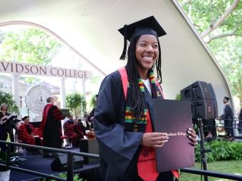 a young Black woman holds a diploma while wearing graduation robes
