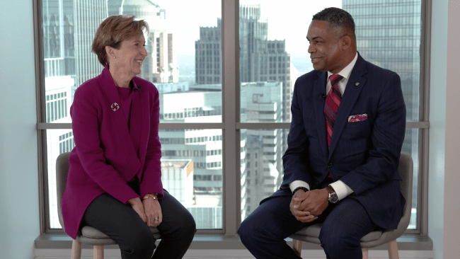 a middle aged white woman and older Black man sit on stools while talking to each other in front of a city skyline