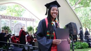 a young Black woman holds a diploma while wearing graduation robes
