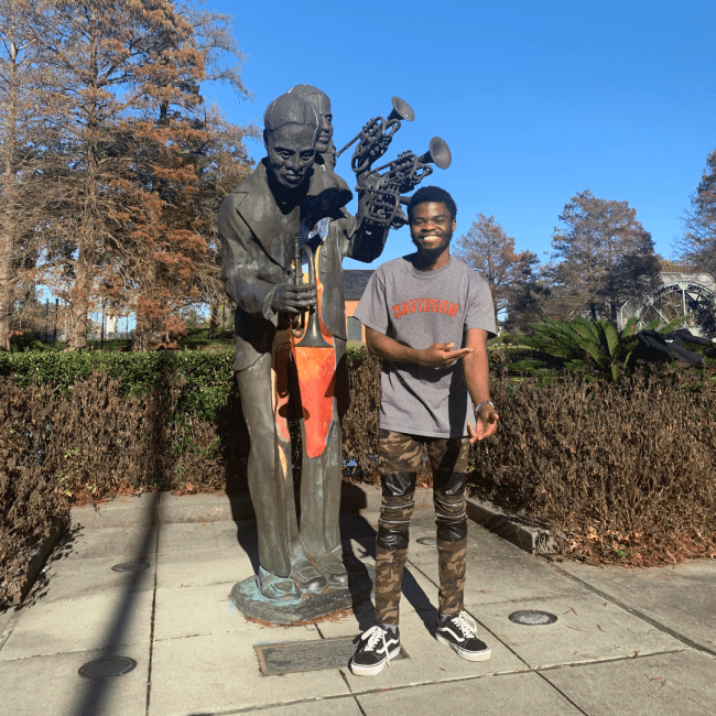 a young Black man wearing a "Davidson" shirt standing next to a Jazz statue