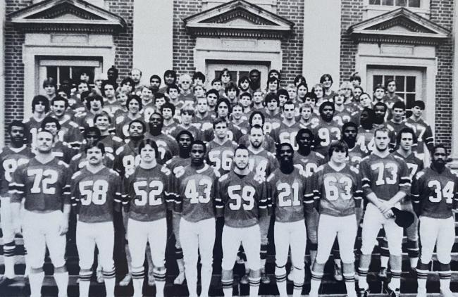 a black and white photo of a football team wearing uniforms and standing in front of a brick building