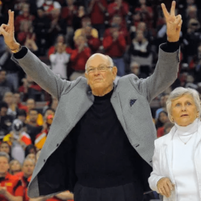 an older white man wears a jacket and gives two peace signs while smiling on a basketball court