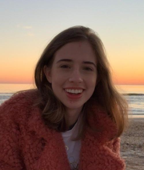 a young white woman with brown hair smiling on a beach with a sunset behind her