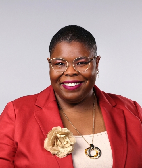a Black woman wearing glasses, a red blazer and gold jewelry on a grey background