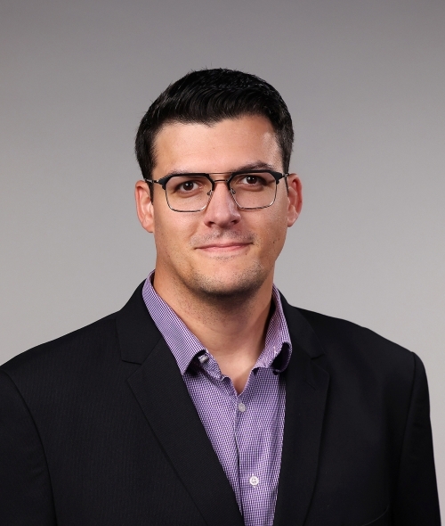 a man with short dark hair wearing glasses, a purple collared shirt and a black blazer on a grey background