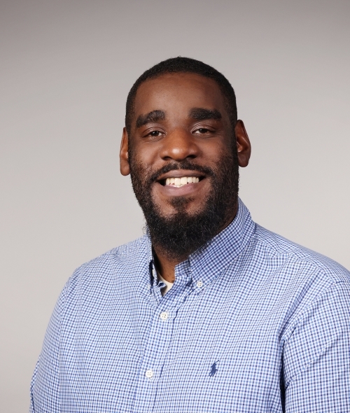 a man with dark facial hair wearing a blue collared shirt on a grey background