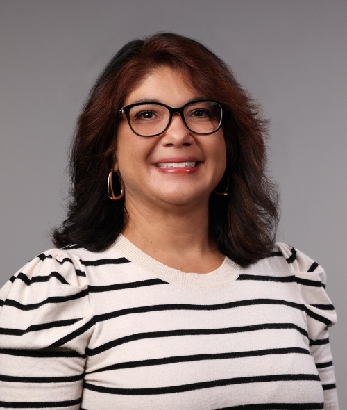 a woman with dark curly hair wearing glasses and a striped top on a grey background