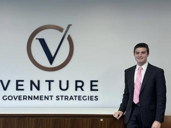 a young white man wears a suit and tie while standing in front of a wall that reads "Venture Government Strategies"