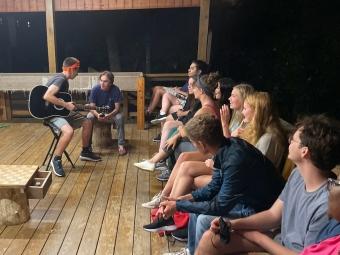 a group of students sit around a cabin as one plays the guitar