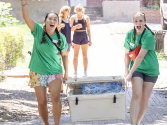 two young women in green shirts carry a bin while smiling and waving