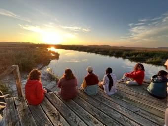 a group of students sit on a dock overlooking marsh at sunset
