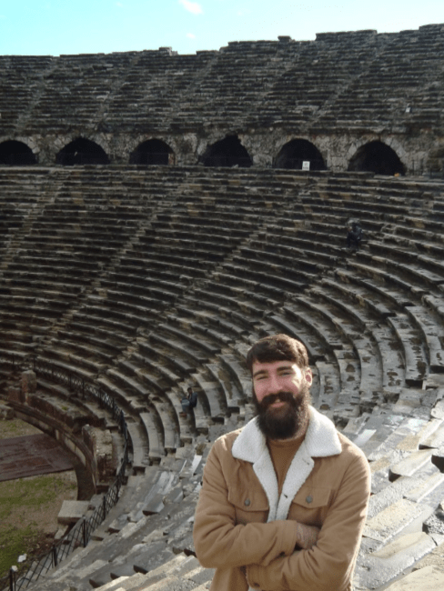 a young man stands in front of an ancient amphitheater