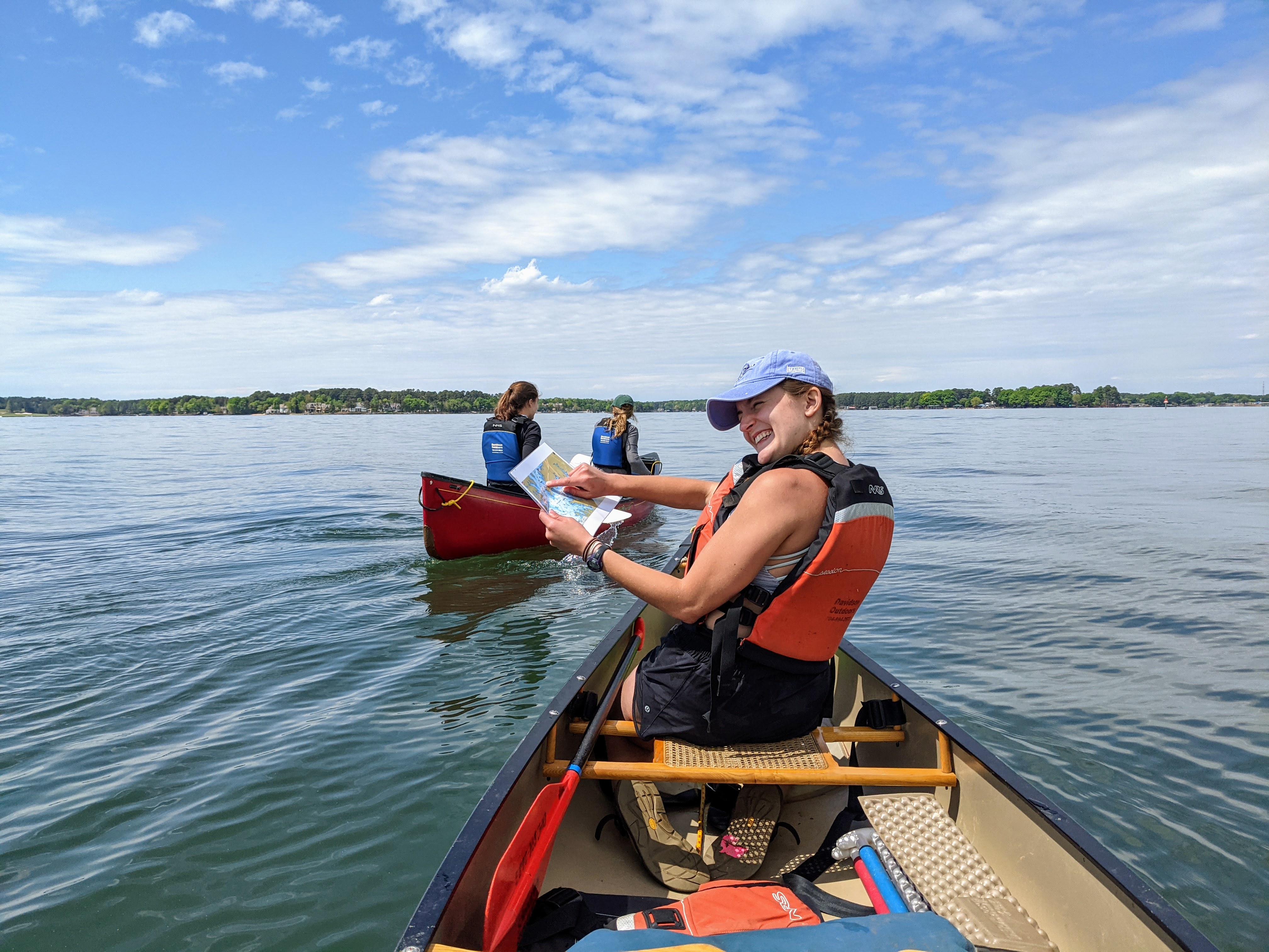 A smiling student in a life jacket holds a map while canoeing on a lake, with two other paddlers in a red canoe nearby