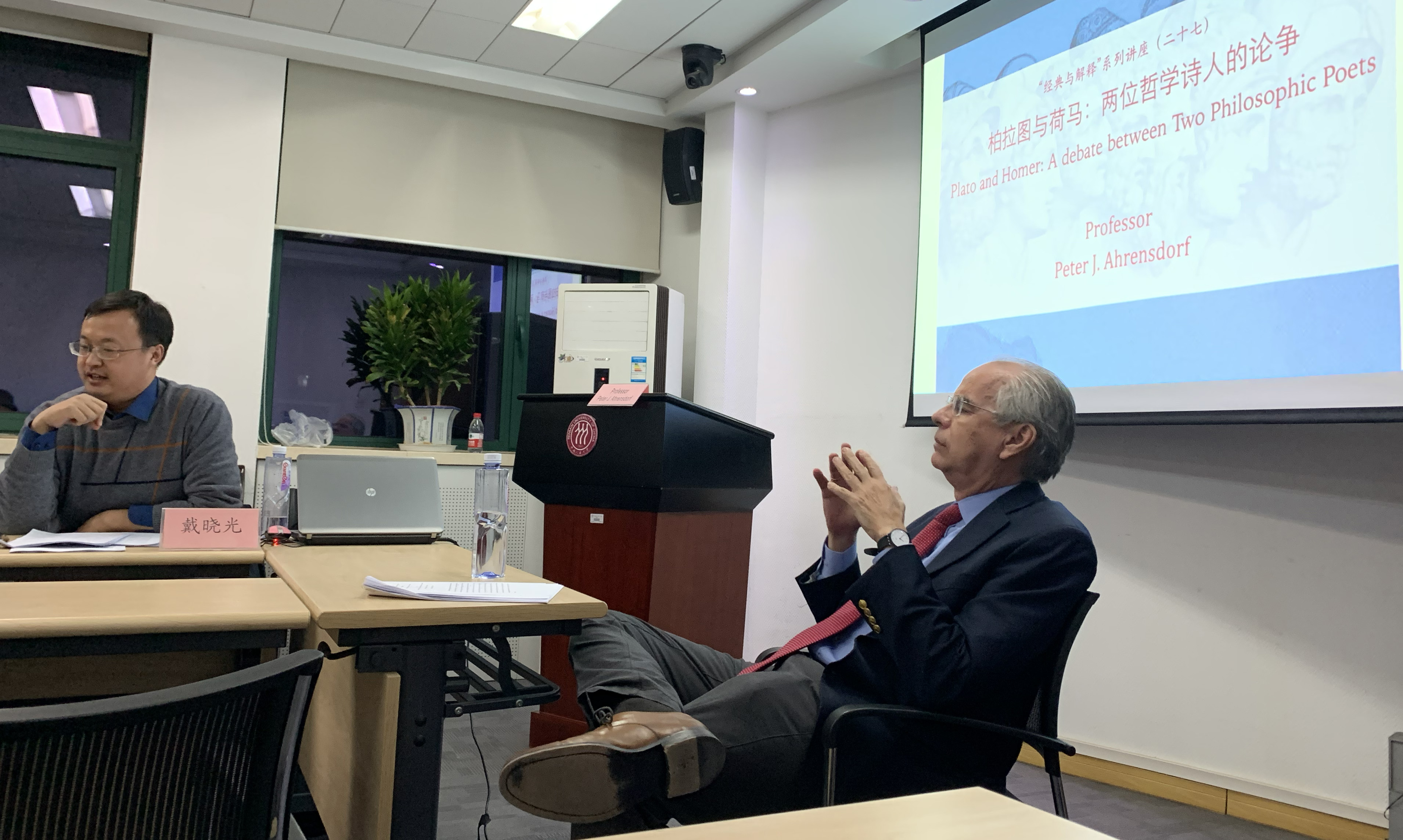 Professor lecturing at a table with powerpoint behind him