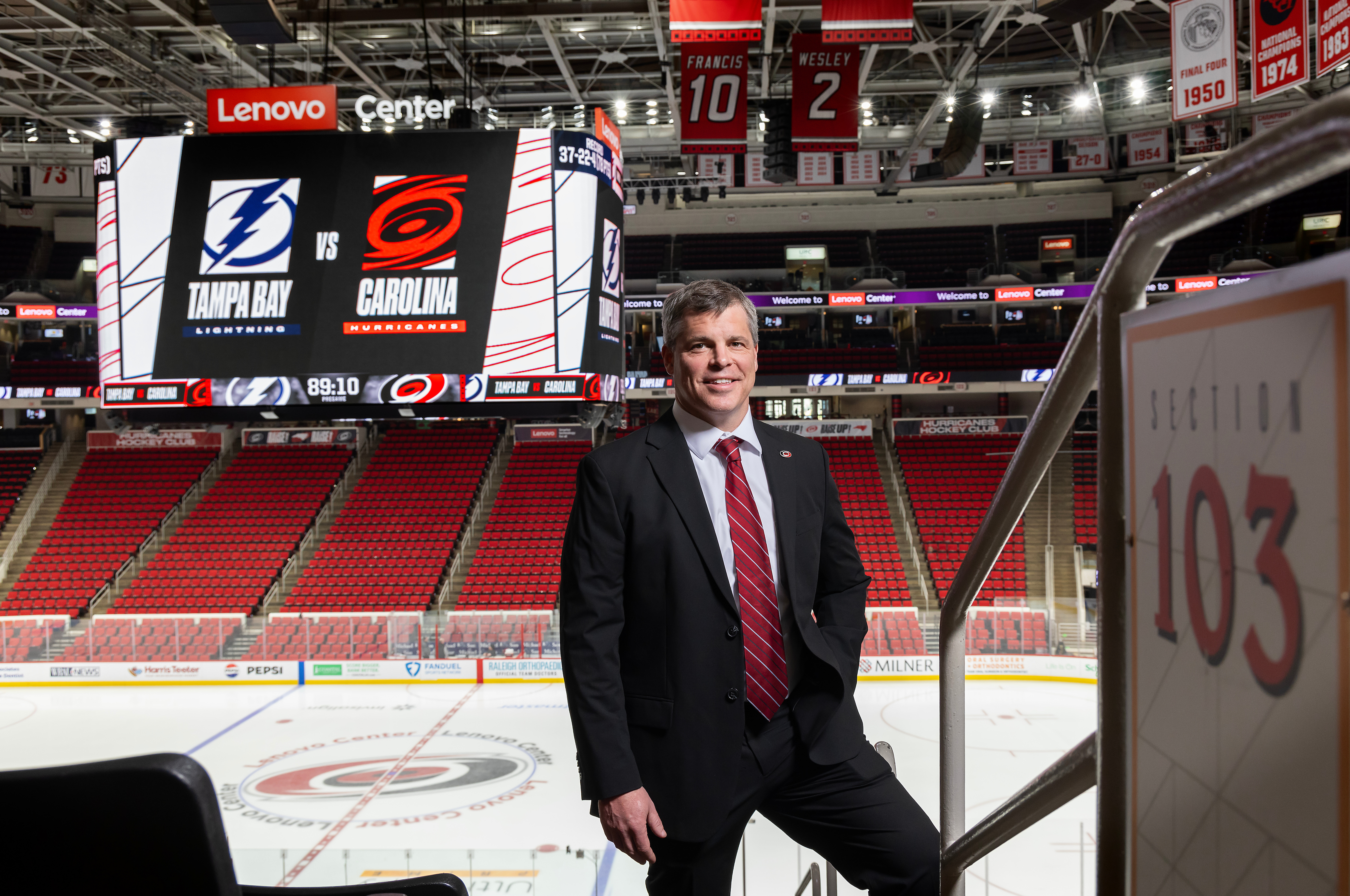 a middle aged white man wearing a black suit with a red tie in front of the Carolina Hurricanes hockey rink