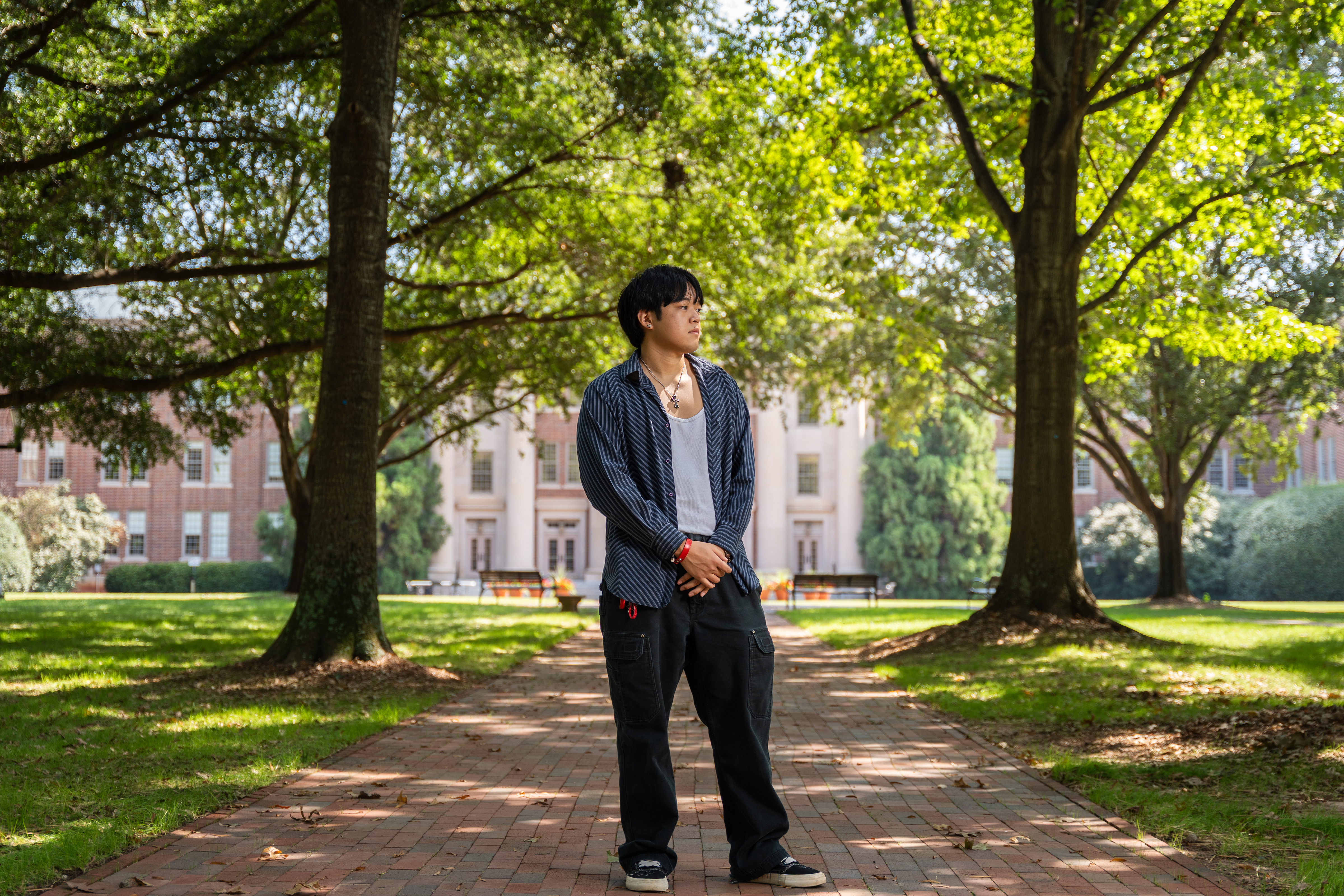 a young man stands on a college campus looking to his side in front of a large building surrounded by trees