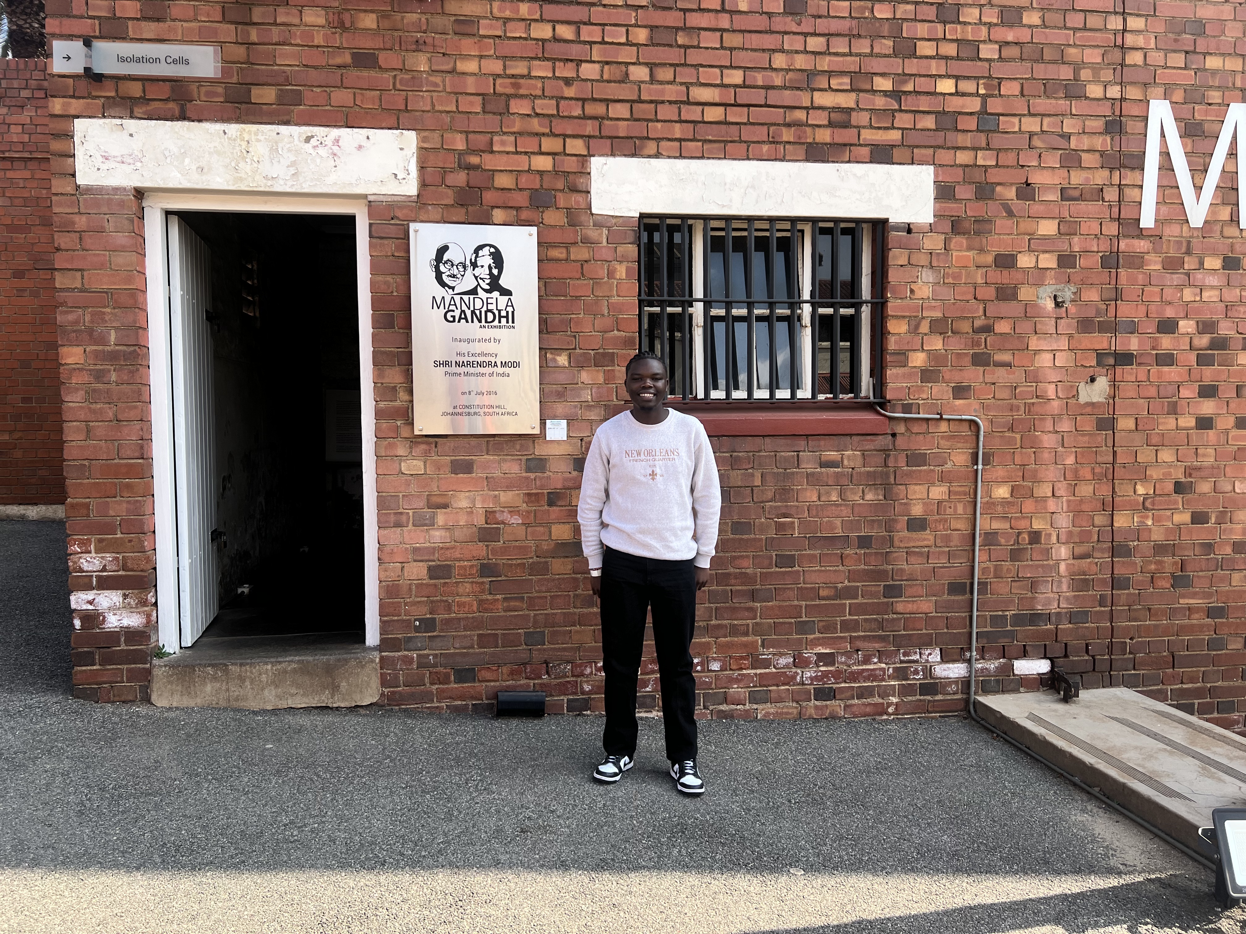 A man in a light grey sweatshirt and black pants stands in front of a red brick building at Constitution Hill in Johannesburg, next to an open doorway labeled "Isolation Cells" and a plaque commemorating the "Mandela Gandhi" exhibition.