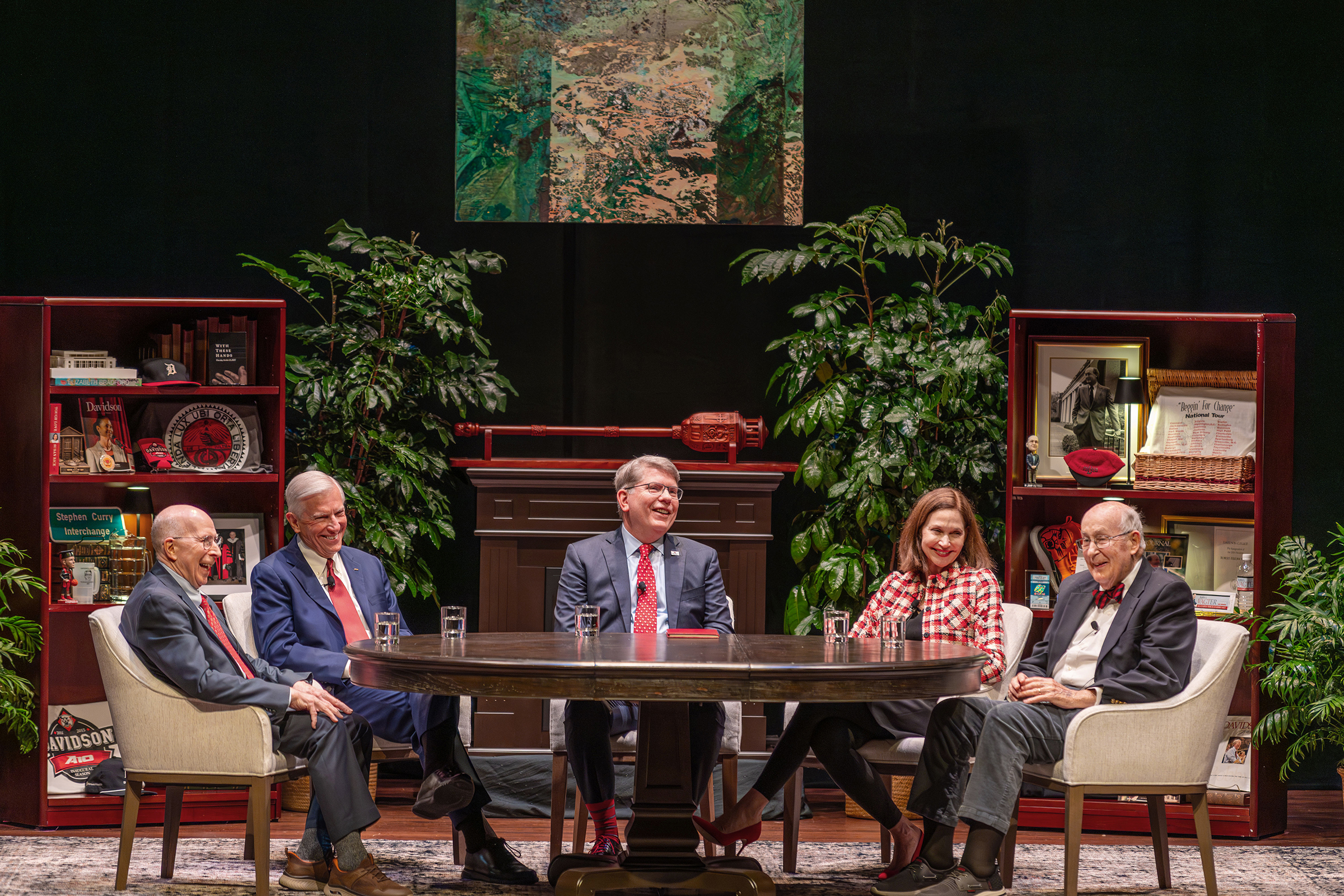 Five people sit around a large oval table on a stage set designed to look like a study, featuring bookshelves filled with memorabilia, potted plants, and an abstract painting hanging in the center.
