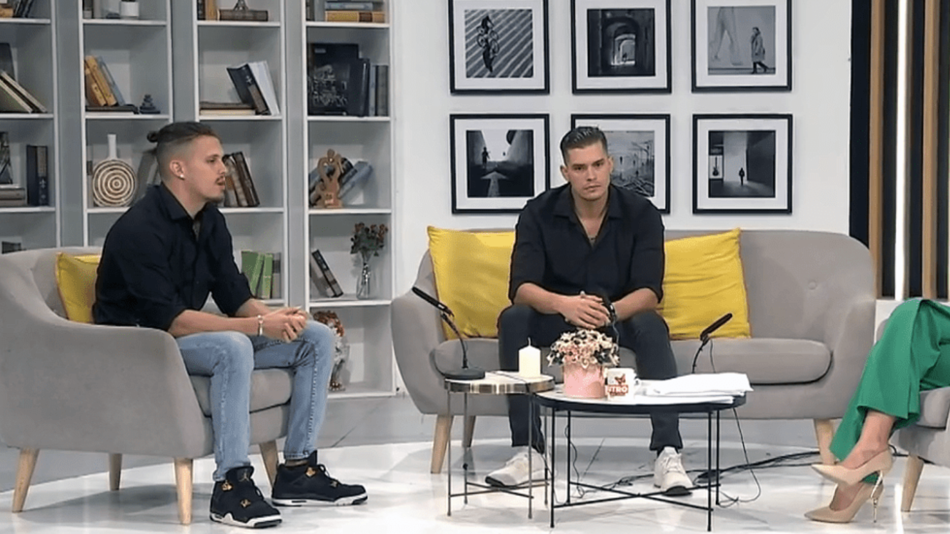 Two men in black shirts sit on a grey sofa with yellow pillows in a studio setting featuring bookshelves and framed wall art.