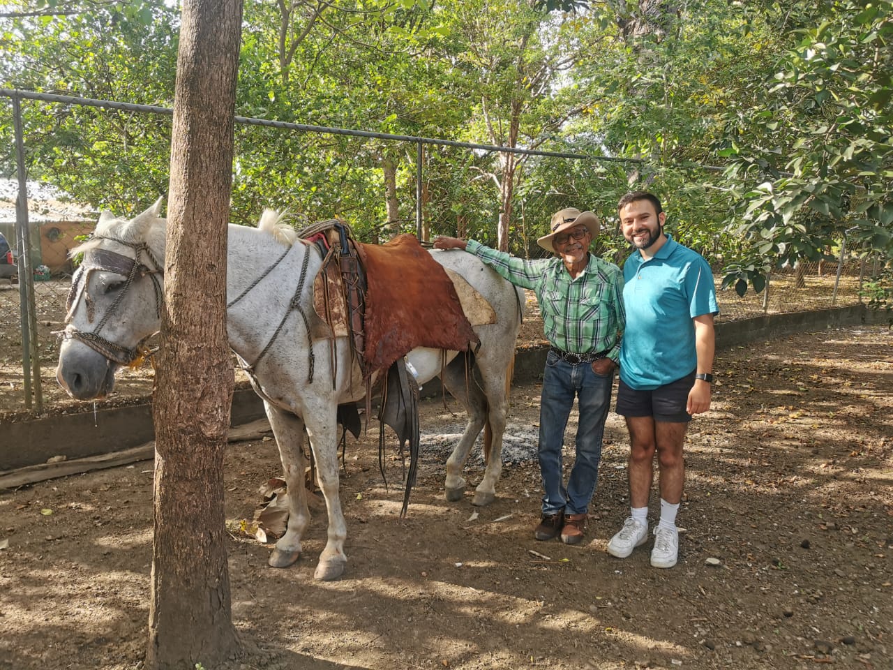 A man in a teal polo and an older man in a cowboy hat and flannel shirt stand together next to a saddled white horse in a fenced, sun-dappled yard.