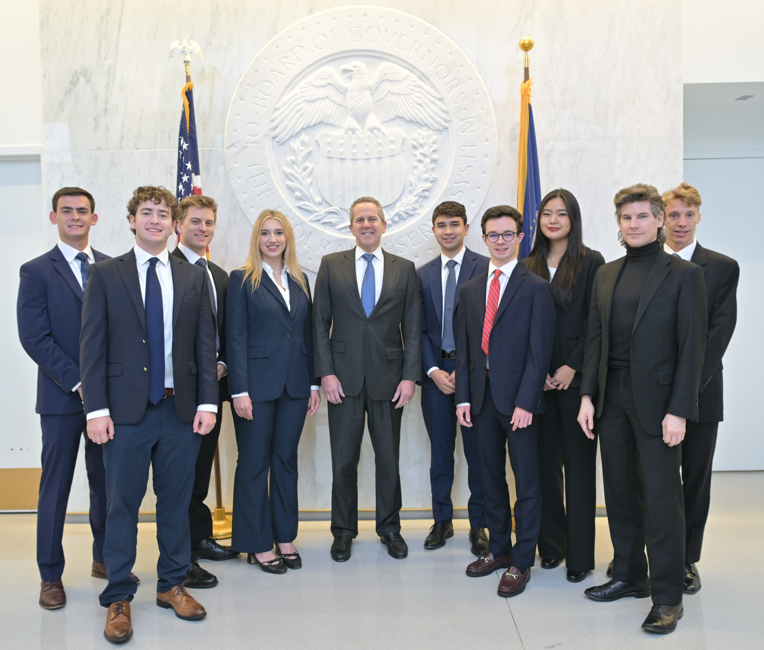 A group of ten professionally dressed students and a central official pose for a formal photo in front of a large, white architectural seal of the Federal Reserve Board of Governors.