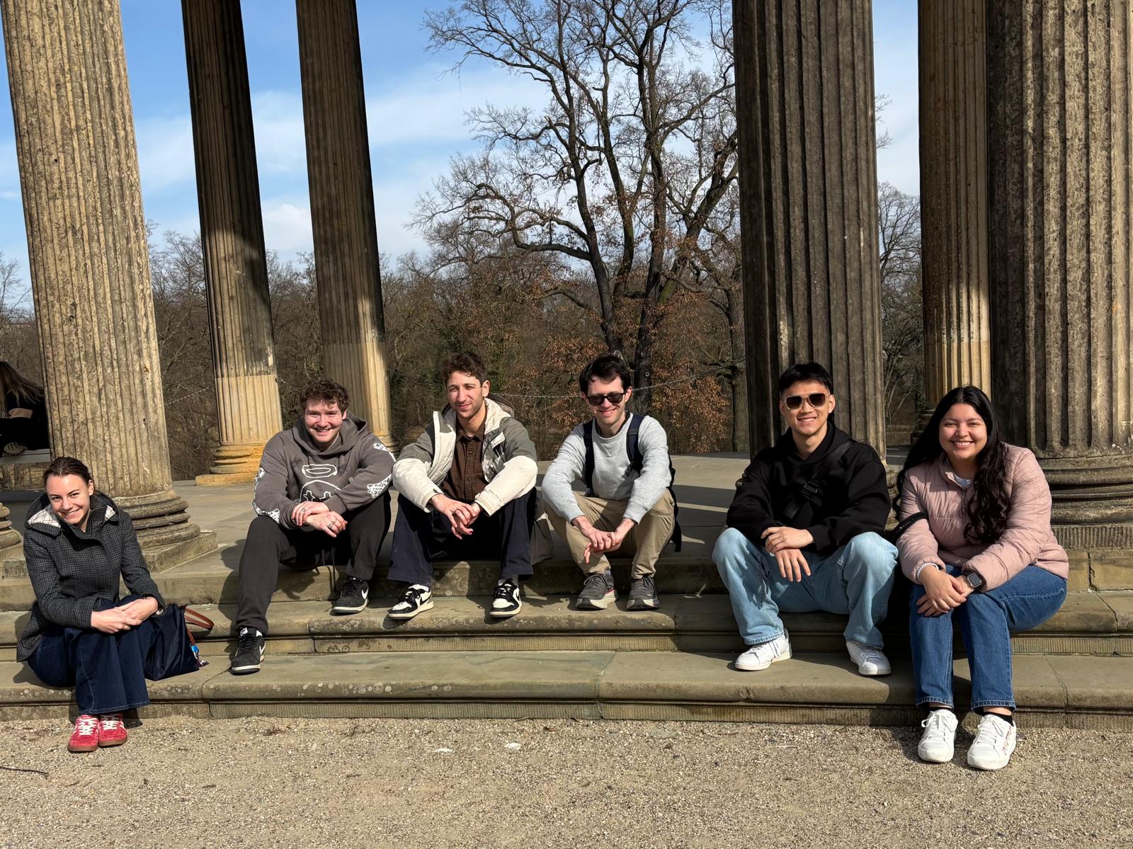 Six individuals sit smiling on the stone steps of a classical pavilion featuring large, fluted columns that frame a view of leafless trees under a clear blue sky.
