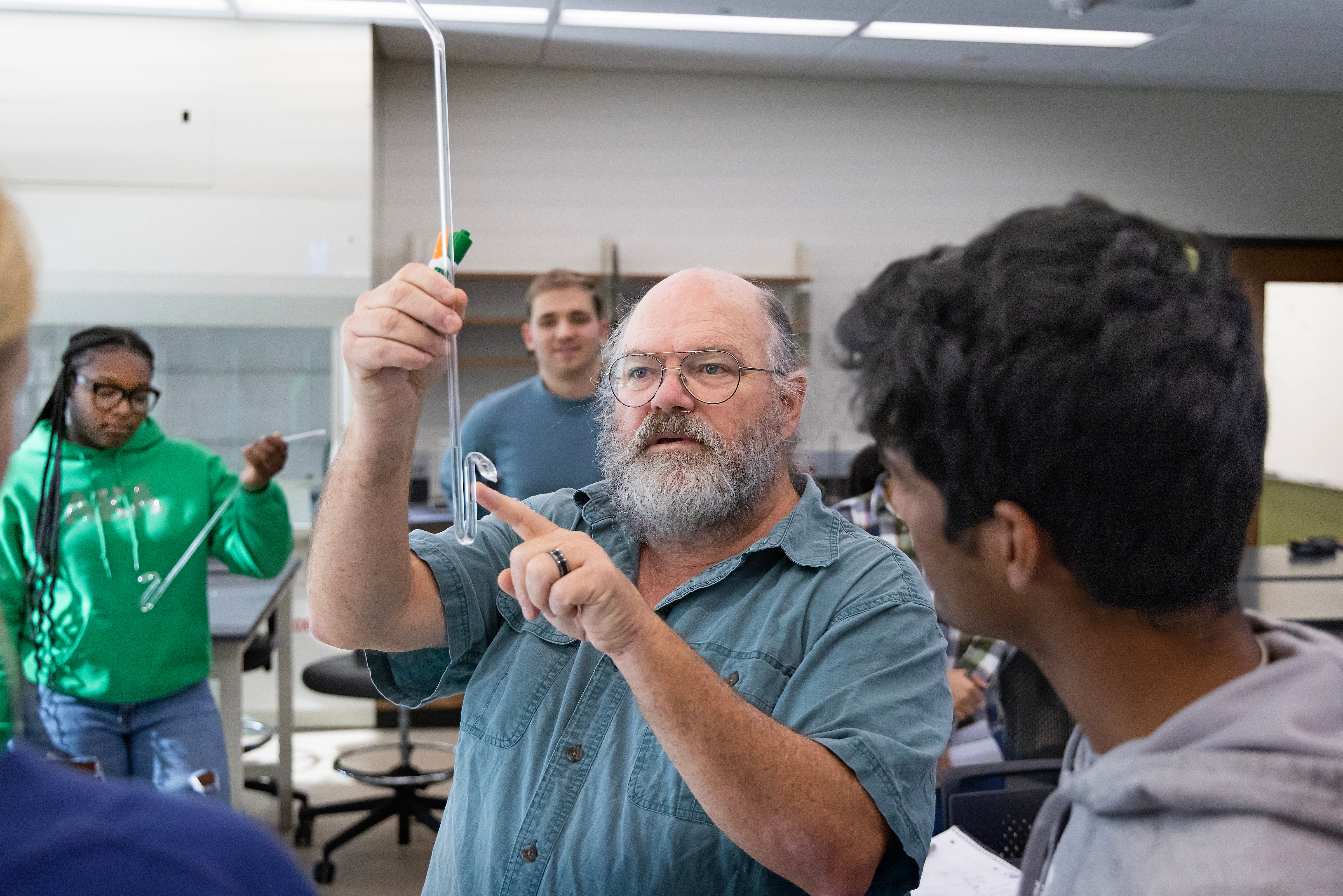 Professor Durwin Striplin, a bearded man wearing glasses, points to a piece of glass laboratory equipment while instructing a student in a chemistry lab setting.