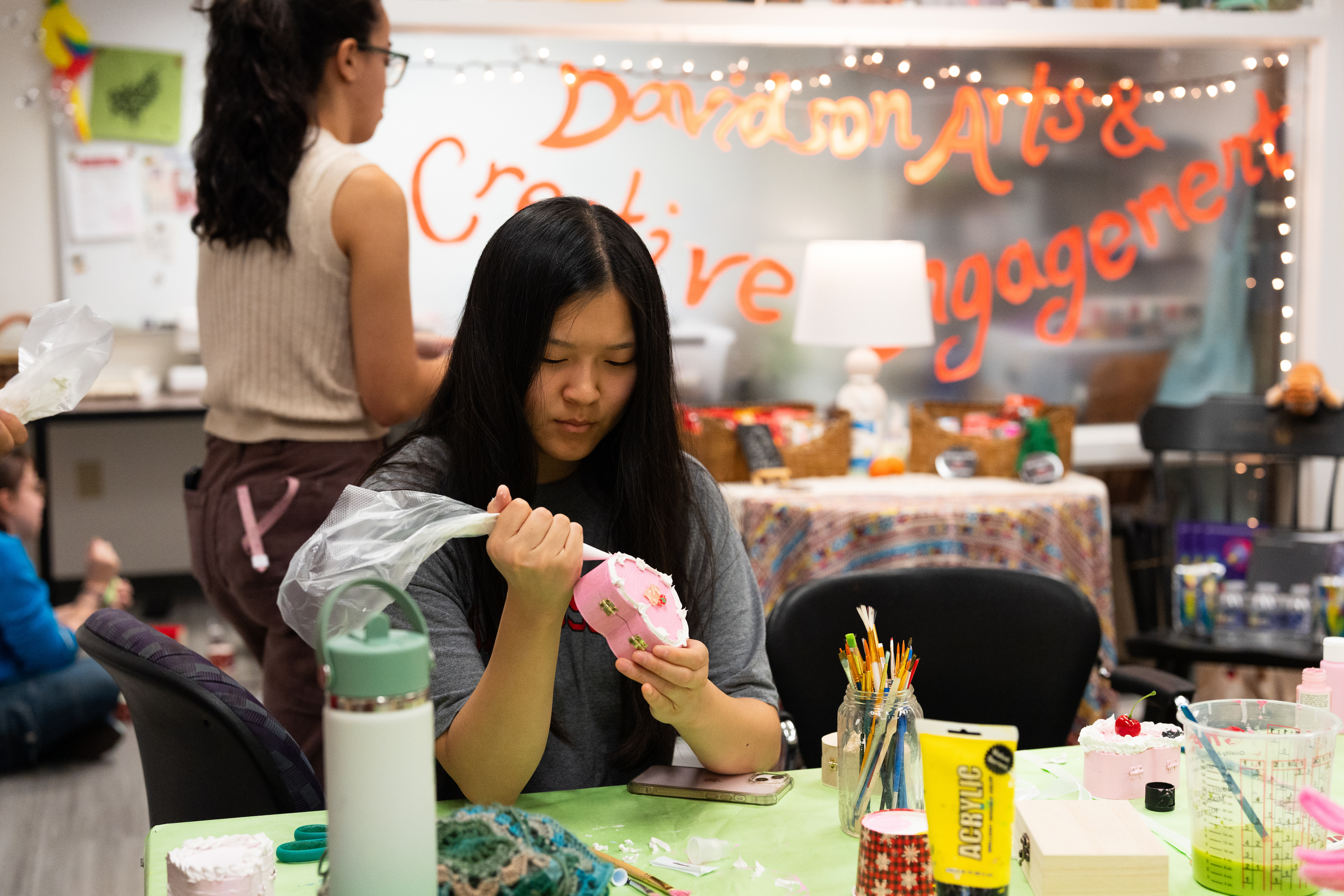 A student focused on her work uses a piping bag to apply white decorative frosting to a small heart-shaped pink jewelry box in a creative studio labeled "Davidson Arts & Creative Engagement."
