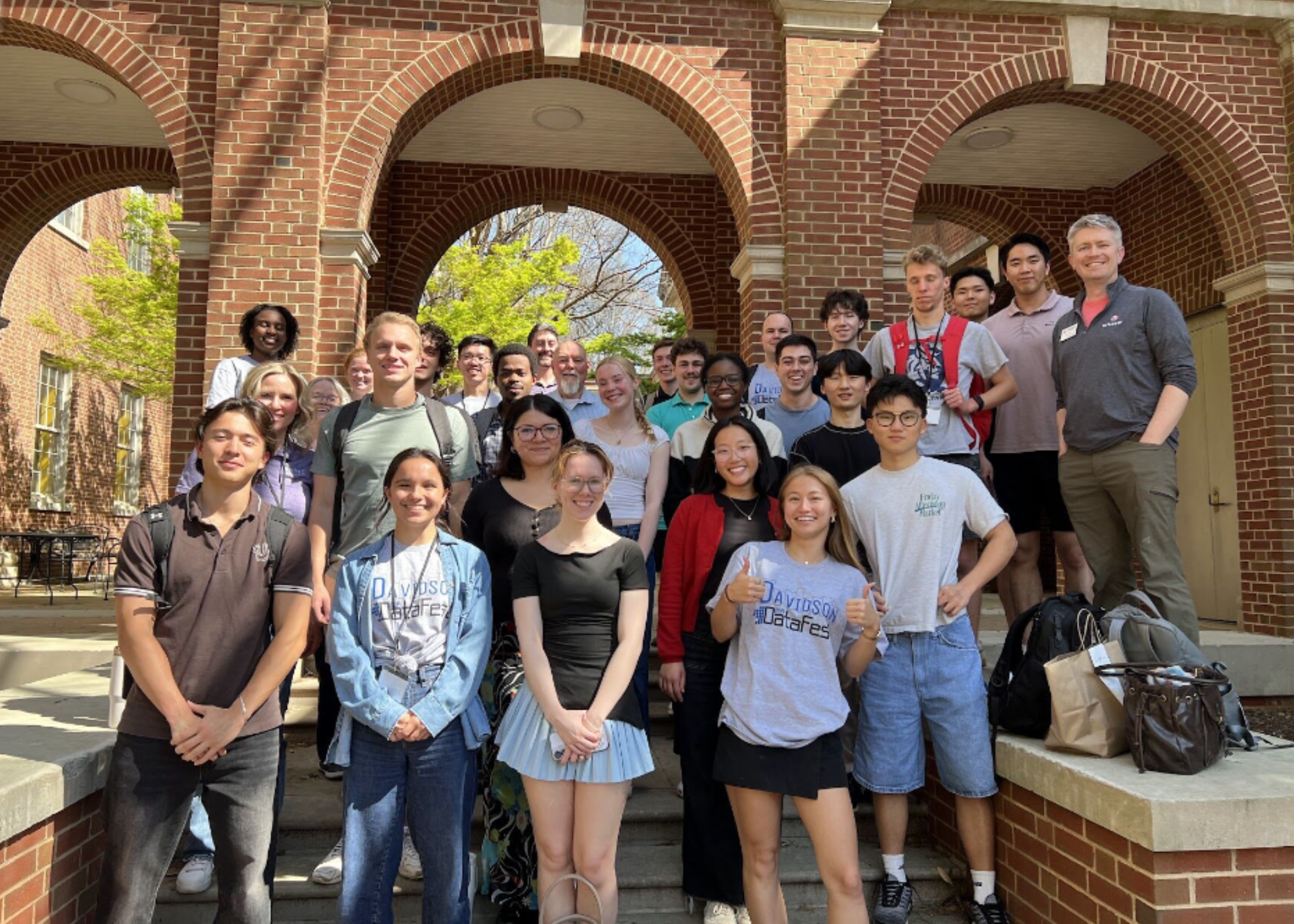A large, diverse group of students and faculty pose together for a commemorative photo on the steps of a brick building with prominent arched entryways.
