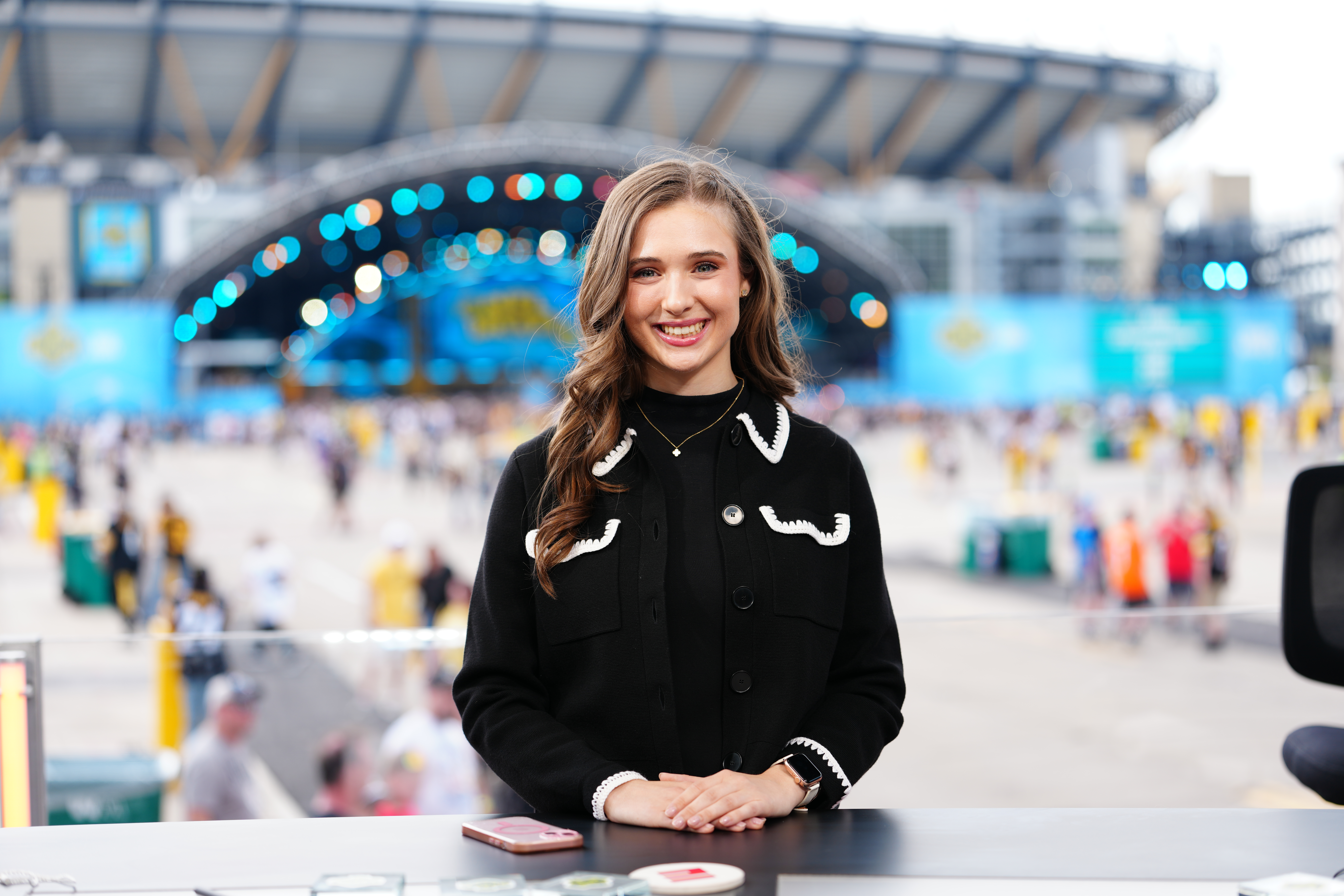 Courtney smiles at a desk with a stadium and large outdoor event stage blurred in the background.