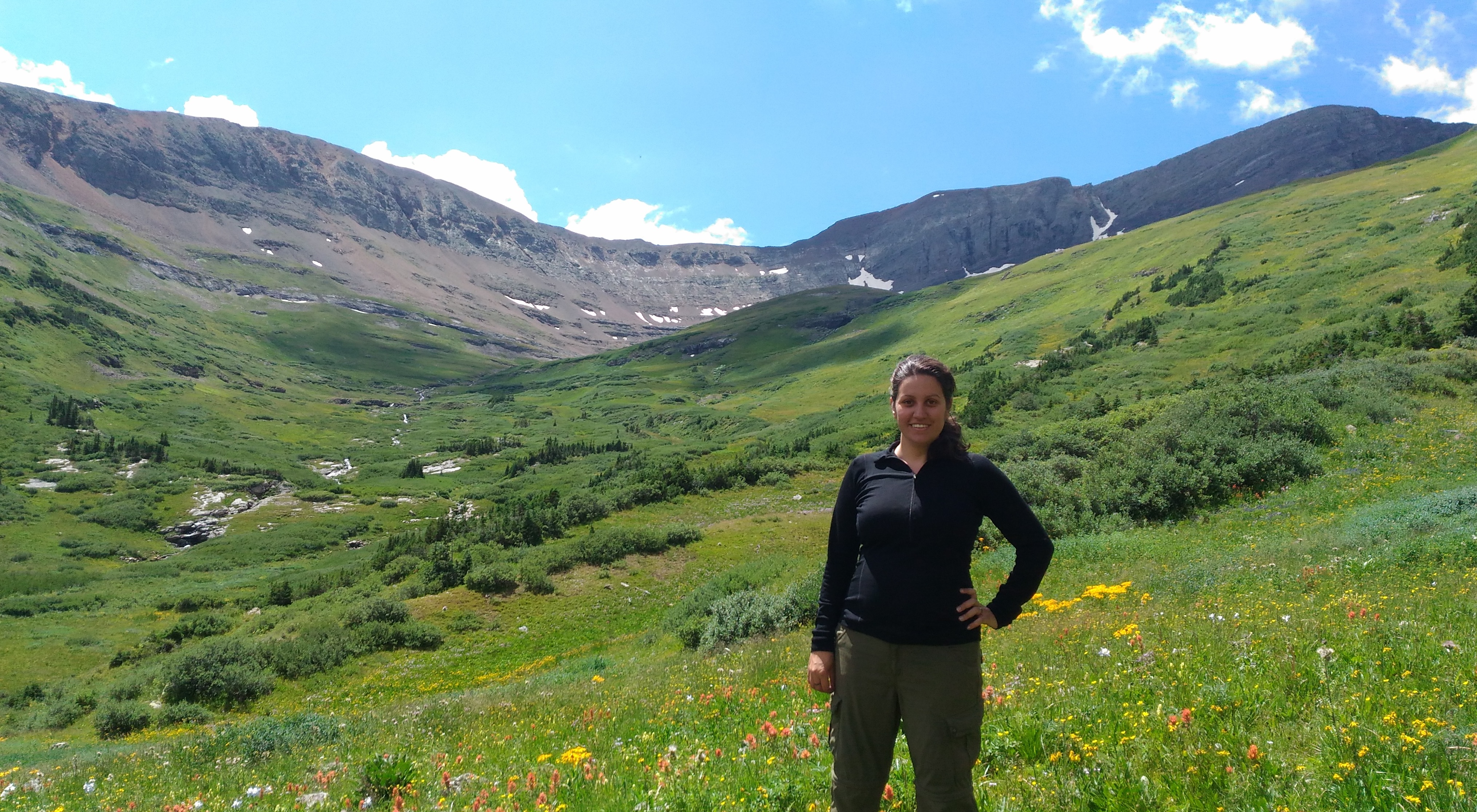 A smiling woman stands in a lush, green mountain valley filled with yellow and red wildflowers under a blue sky with light clouds.