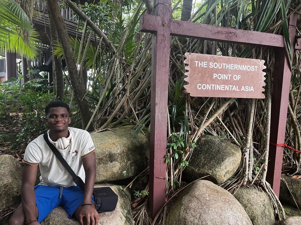A young man sits on large rocks next to a wooden signpost reading "The Southernmost Point of Continental Asia," surrounded by tropical foliage.