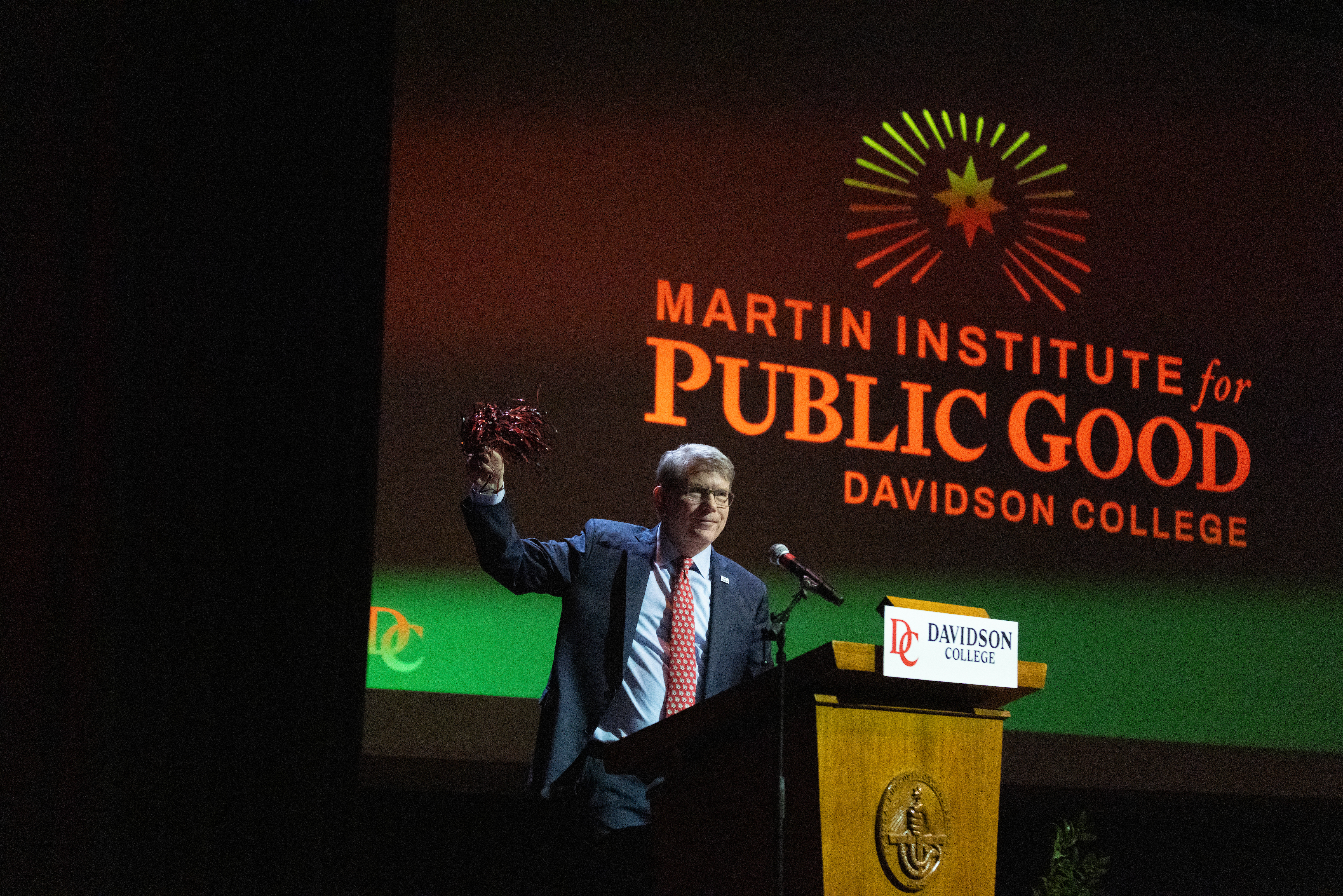 A man in a suit stands at a Davidson College podium on a dimly lit stage, raising a pom-pom in one hand beneath a large projection screen that reads "Martin Institute for Public Good."