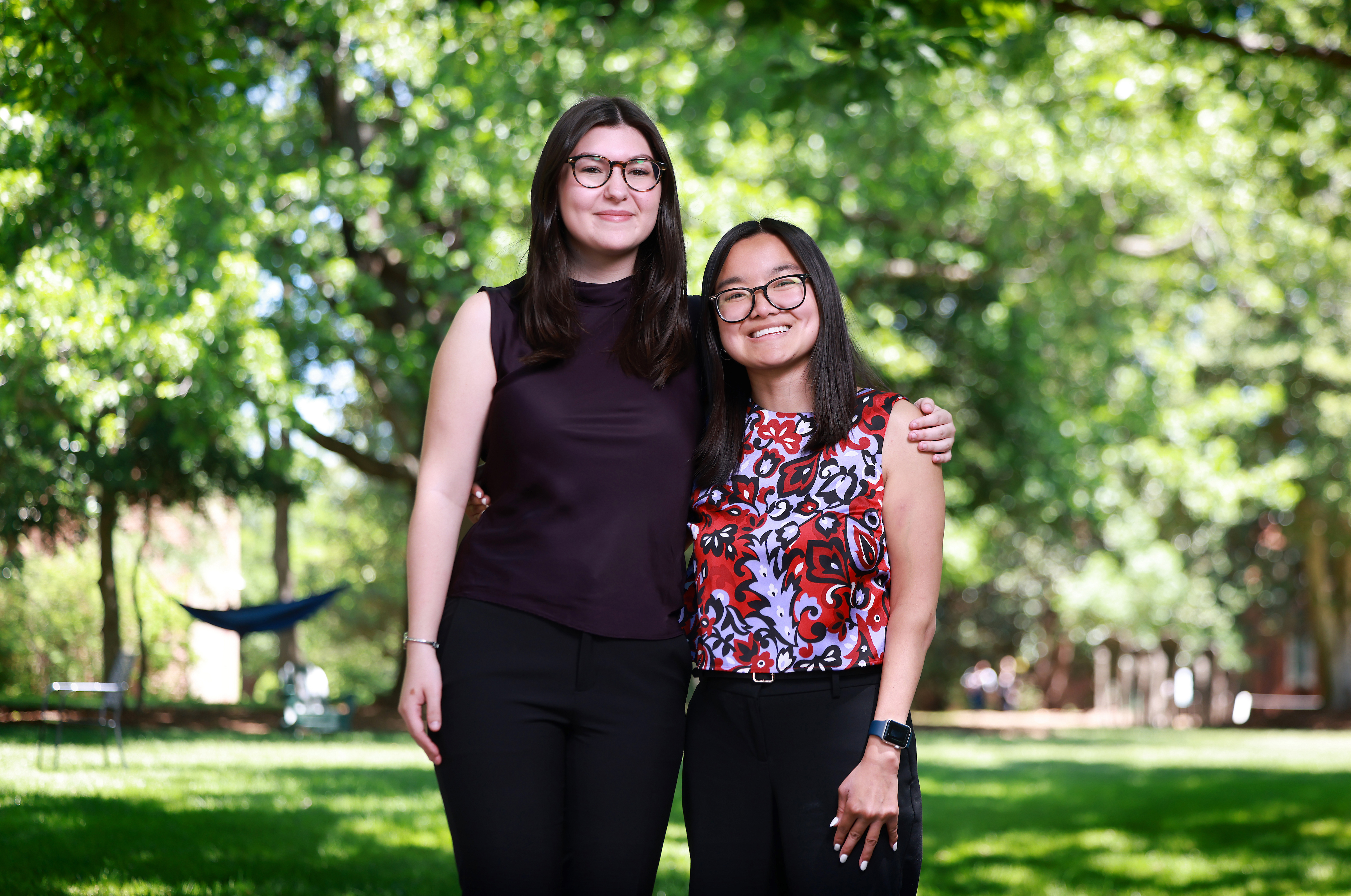 Two smiling students wearing glasses stand side-by-side with arms around each other on a sunny, green campus lawn surrounded by trees.
