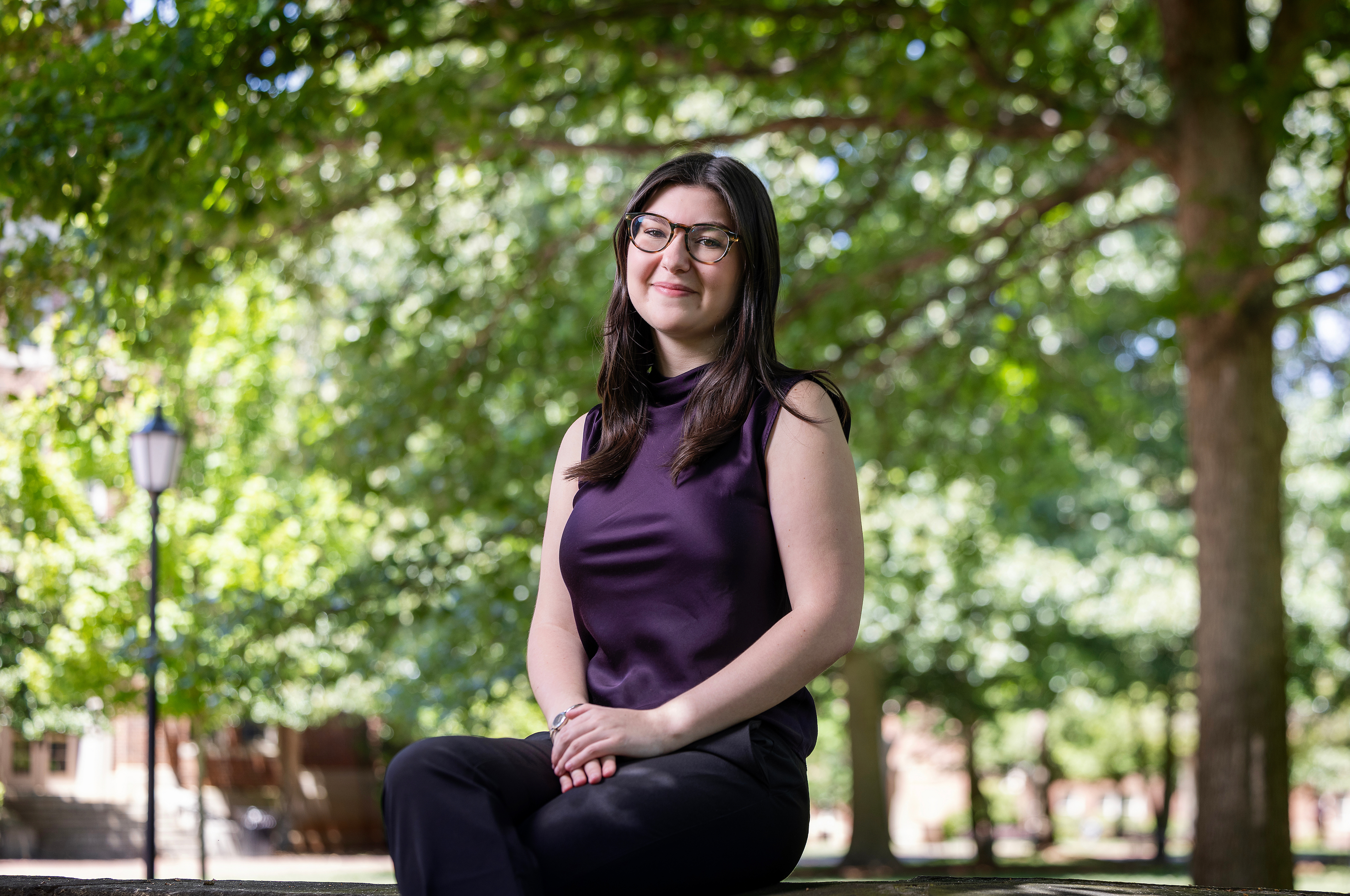 A smiling student wearing glasses and a dark purple sleeveless top sits outside on a stone ledge beneath leafy green trees.