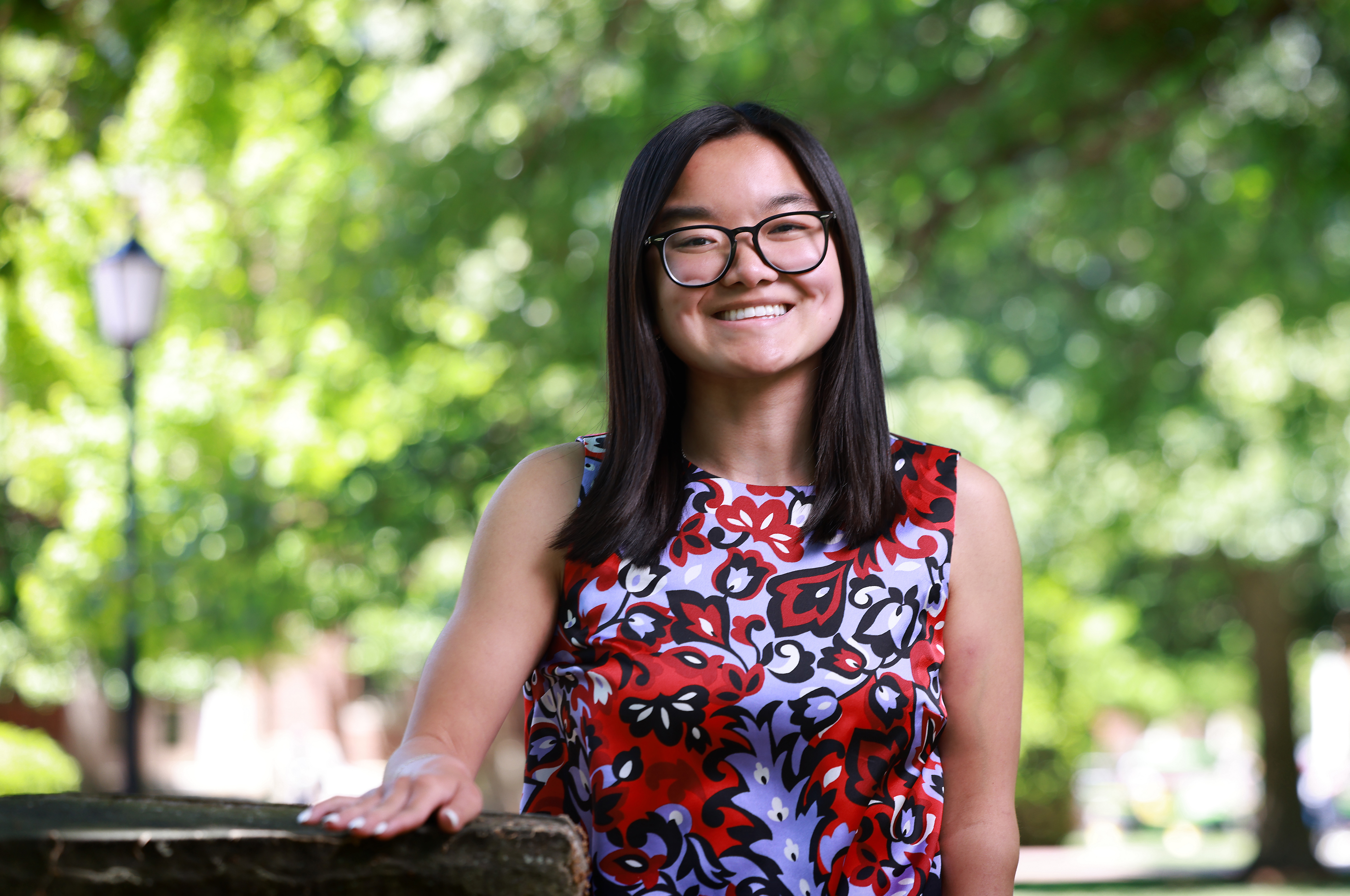 A smiling student with glasses and straight, dark hair poses outside wearing a sleeveless floral patterned blouse.