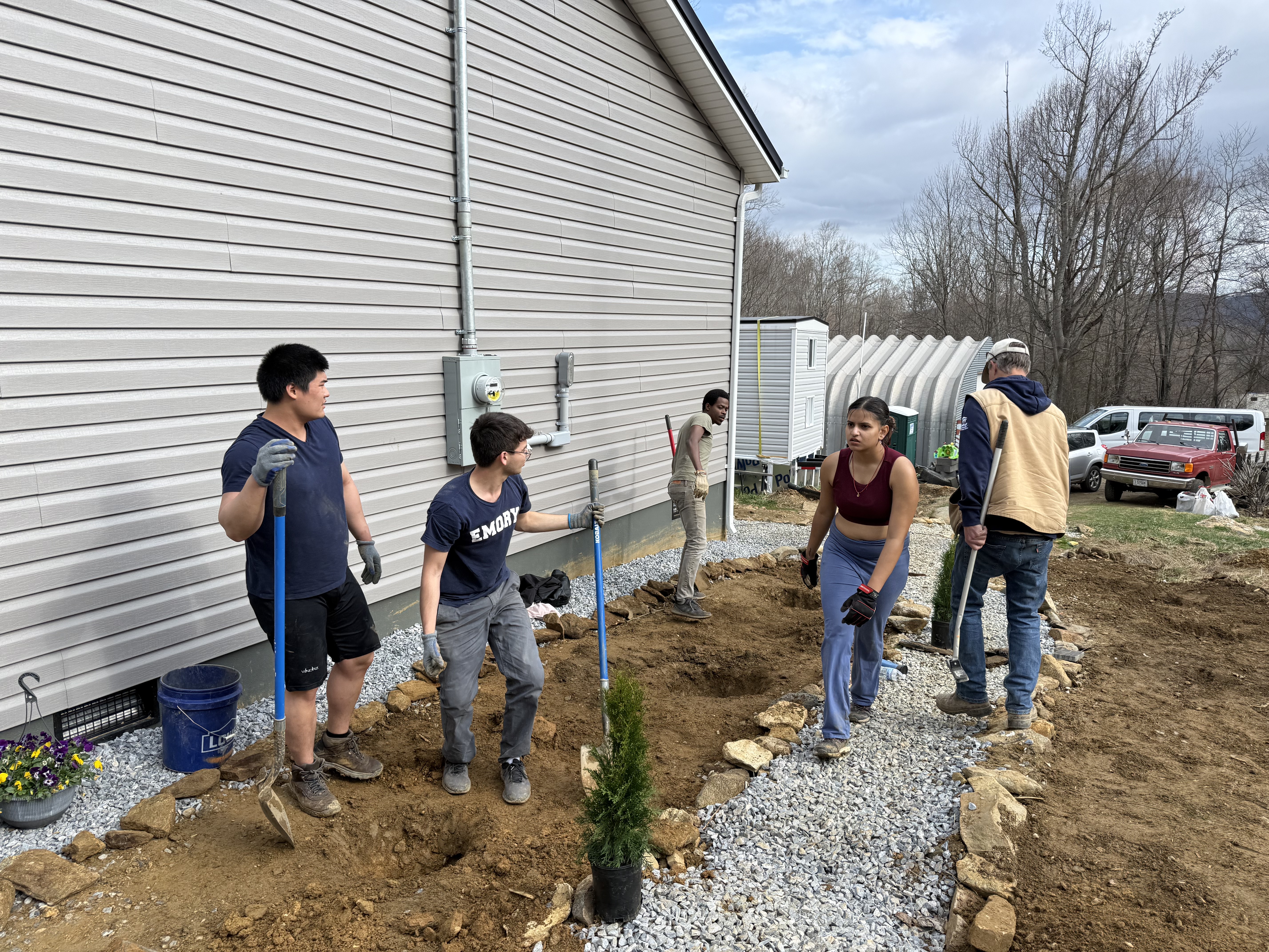 A group of volunteers works together on an outdoor landscaping project next to a house, digging trenches and laying gravel and stone borders.