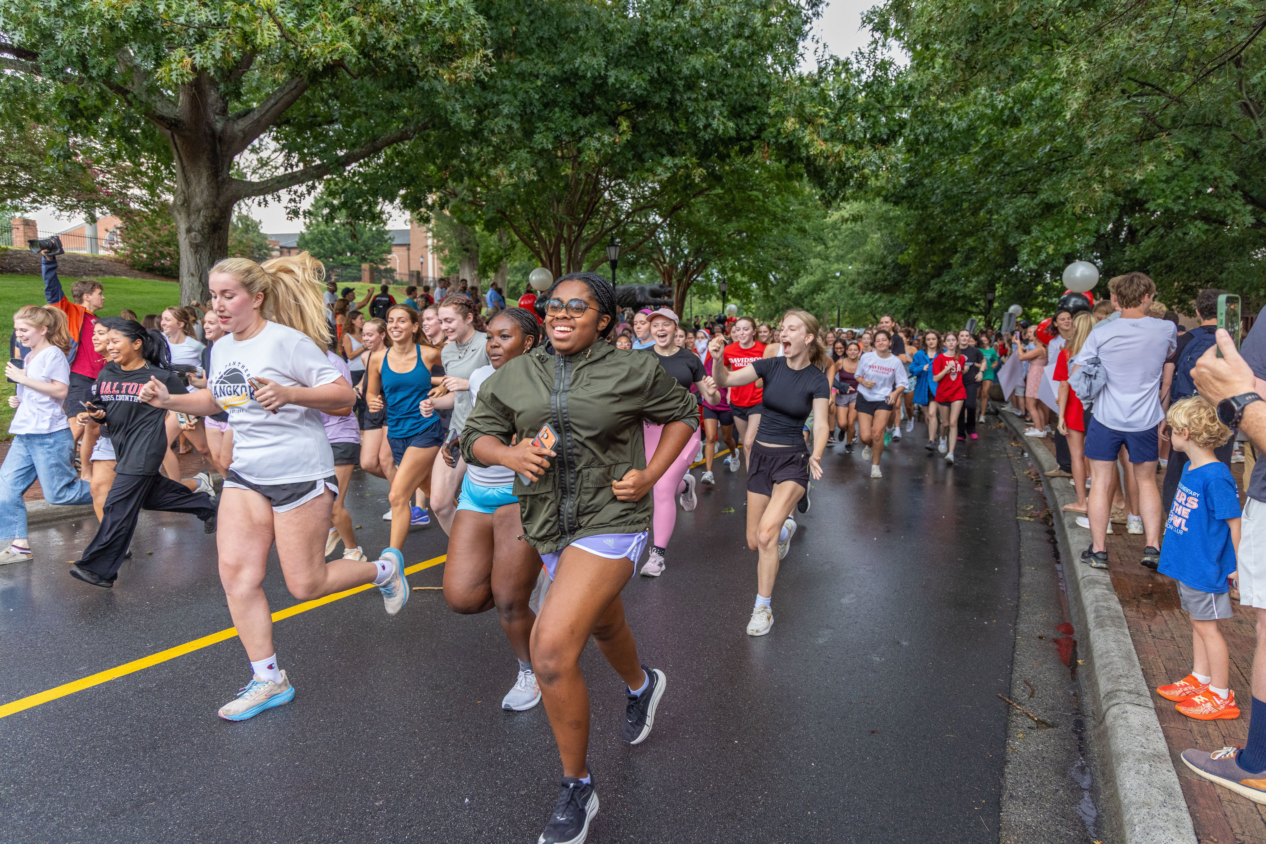 A large group of smiling young runners race down a tree-lined paved road while spectators cheer along the sidewalk.