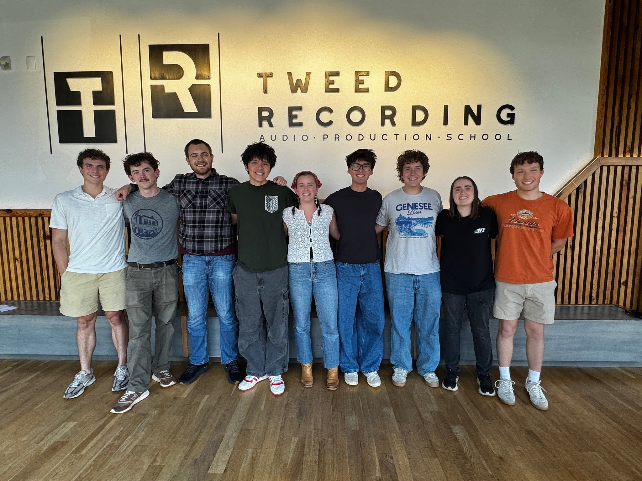 A group of eight young adults stands in a line with their arms around each other's shoulders, posing for a photo inside a modern room in front of a wall with a logo that reads "Tweed Recording Audio Production School."
