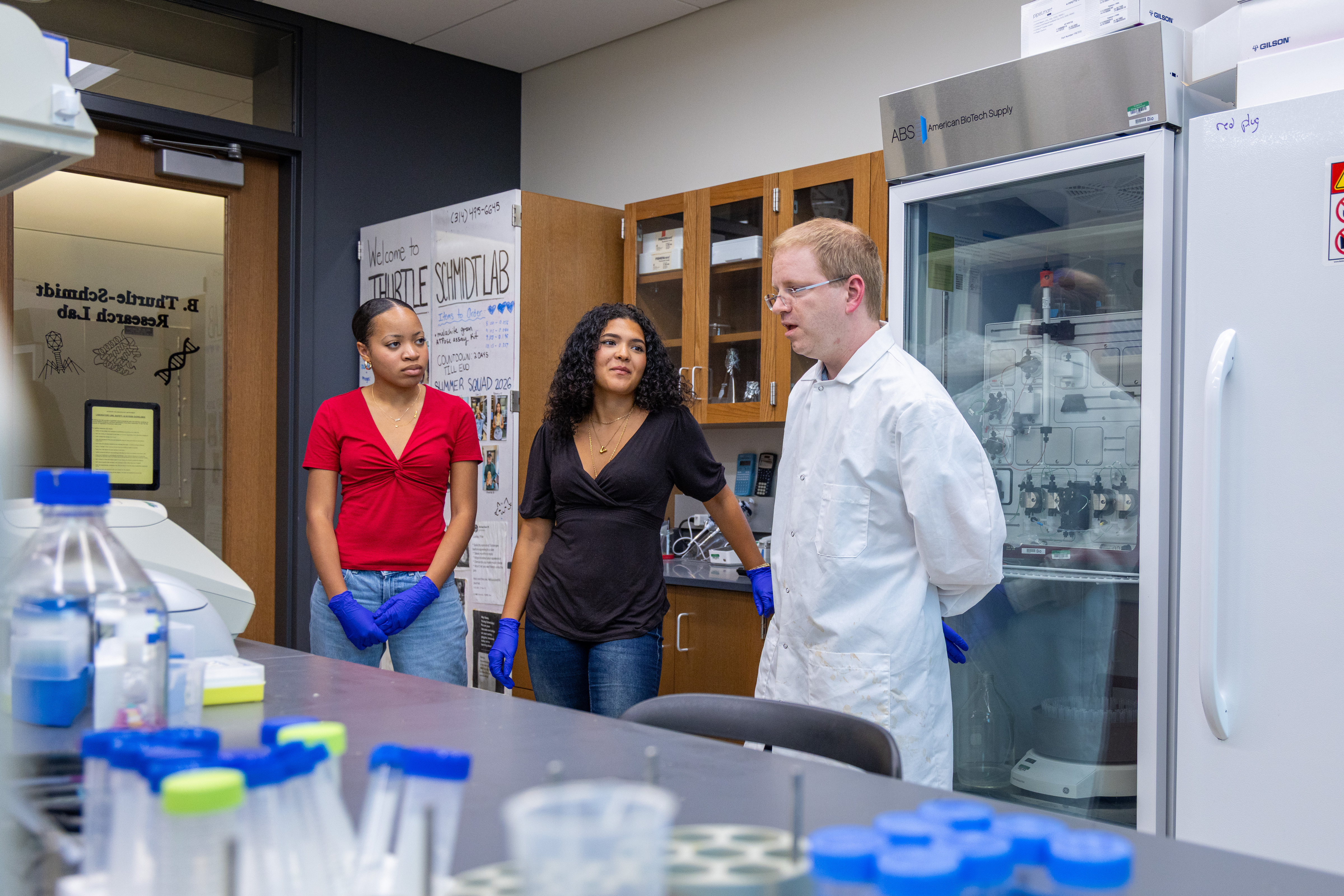Dr. Thurtle-Schmidt, wearing a white lab coat, speaks with two student researchers wearing protective blue gloves inside a science laboratory.