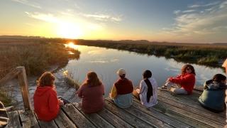 a group of students sit on a dock overlooking marsh at sunset