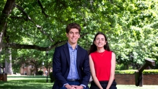 a young man and a young woman sit together on a brick wall in front of green trees