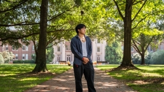 a young man stands on a college campus looking to his side in front of a large building surrounded by trees