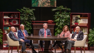 Five people sit around a large oval table on a stage set designed to look like a study, featuring bookshelves filled with memorabilia, potted plants, and an abstract painting hanging in the center.