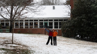 Two people wearing colorful winter jackets stand with arms around each other on a snow-dusted grassy slope in front of a brick building with a snow-covered roof and a large evergreen tree.