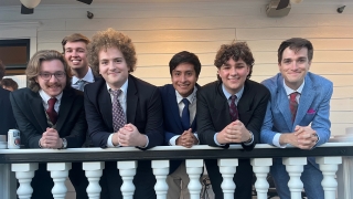 Six young men in formal suits and ties lean against a white balcony railing with classic balusters in front of a light-colored wooden house.