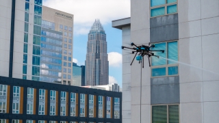 A professional-grade cleaning drone hovers beside a modern apartment building, spraying a stream of water onto the exterior windows against a city skyline featuring the Bank of America Corporate Center in Charlotte.