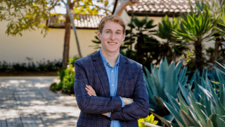 A young white man wearing a blue blazer in a tropical setting, arms crossed