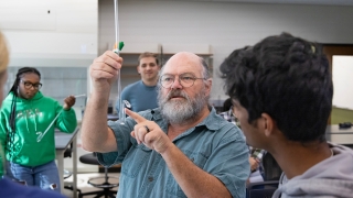 Professor Durwin Striplin, a bearded man wearing glasses, points to a piece of glass laboratory equipment while instructing a student in a chemistry lab setting.