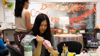 A student focused on her work uses a piping bag to apply white decorative frosting to a small heart-shaped pink jewelry box in a creative studio labeled "Davidson Arts & Creative Engagement."
